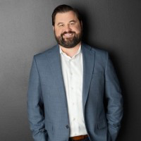 A man with a beard, wearing a blue suit and white shirt, smiles while standing against a dark gray background.