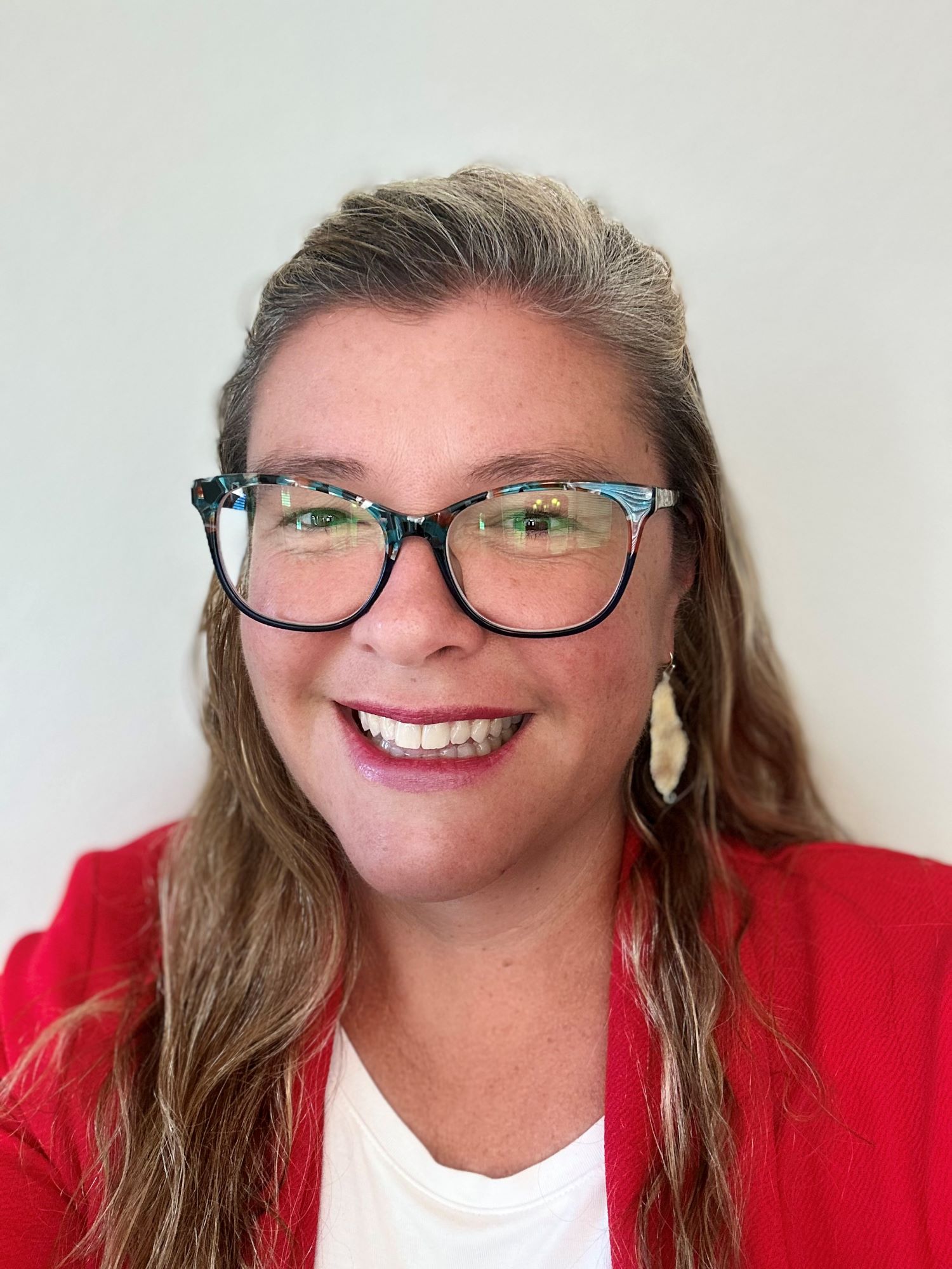 Woman with glasses smiling, wearing a red blazer, white top, and feather-shaped earrings against a white background.