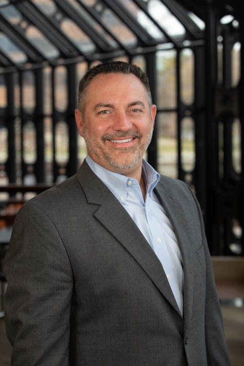 Smiling man in a gray suit jacket and blue shirt stands in a sunlit, windowed atrium.