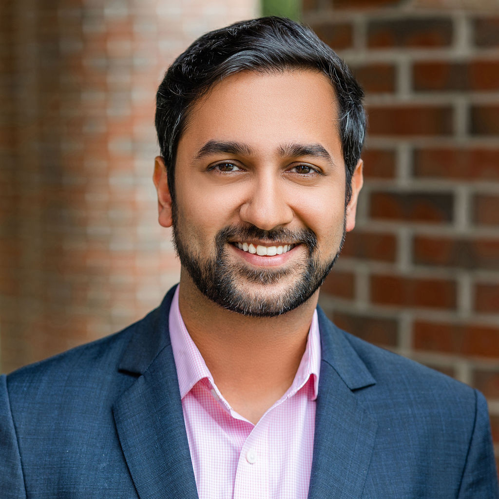 Man in a suit and pink shirt smiling in front of a brick wall background.