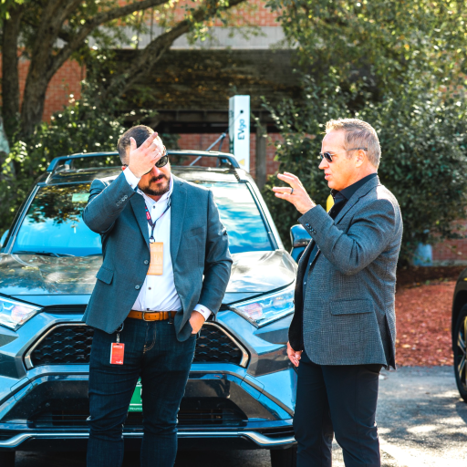 Two men talking in front of a car; one adjusts his glasses, and the other raises his hand in conversation.