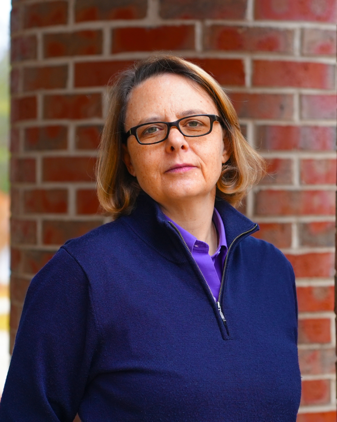 Person with short hair and glasses, wearing a dark sweater, stands in front of a brick column.
