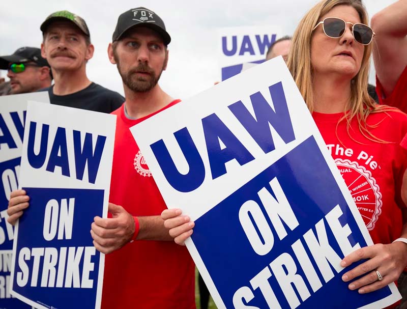 Protesters holding "UAW On Strike" signs, wearing casual clothing, gathered outdoors.