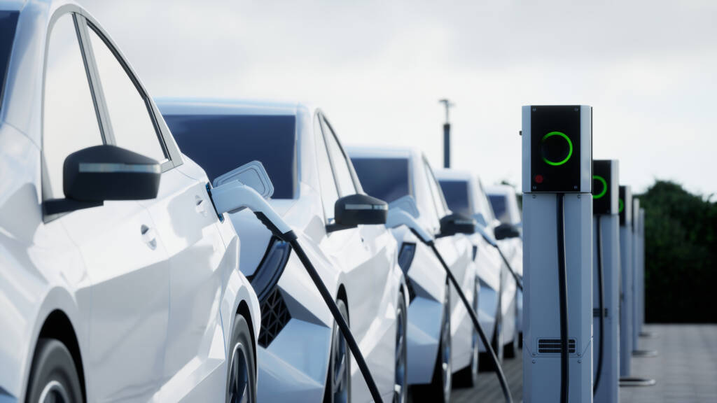 Multiple white electric cars are parked and charging at a line of charging stations outdoors.