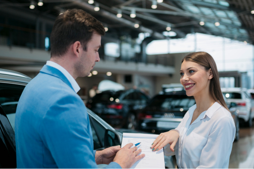 A man and woman discussing over a clipboard in a car showroom, surrounded by vehicles.