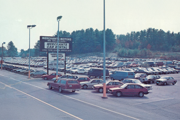 A large parking lot filled with many parked cars and a Merchant Motors sign, surrounded by trees in the background.
