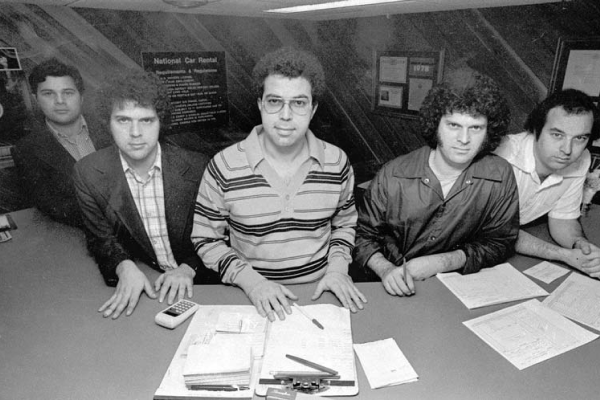 Five men standing behind a desk with paperwork and a calculator.