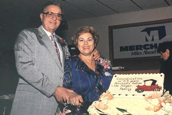 An elderly couple smiles while cutting a decorated cake at a celebration event.