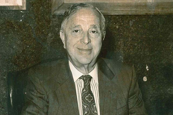 Elderly man in a suit and tie, smiling while seated, with a dark textured background.