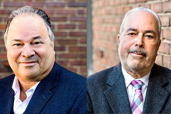 Two men in suits stand in front of a brick wall, smiling slightly at the camera.