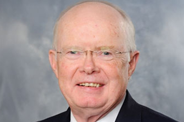A bald man wearing glasses and a suit smiles against a gray background.