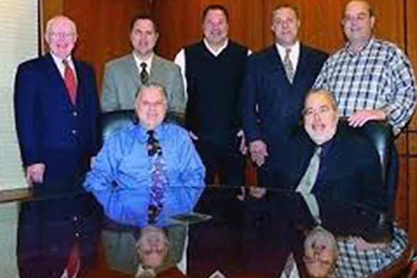 Seven men in business attire pose around a conference table in an office.