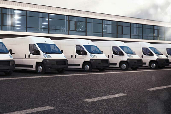 A row of white delivery vans parked in front of a modern glass building under a cloudy sky.