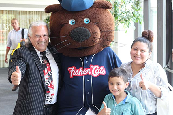 A man, woman, and child smile and give thumbs up beside a mascot in a "Fisher Cats" jersey and hat.