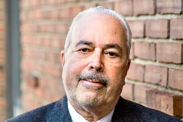 Man with short gray hair and a goatee, wearing a suit, standing against a brick wall.