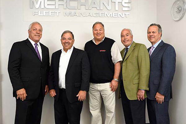 Five men in business attire standing in front of a sign that reads "Merchants Fleet Management.