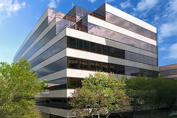 Modern glass office building with angular design, surrounded by trees, under a blue sky with clouds.