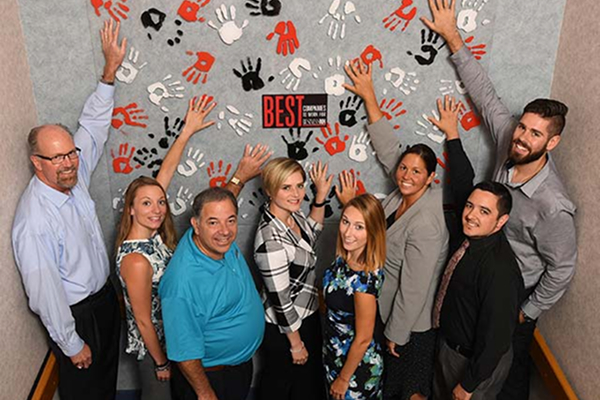 A group of people smiling and touching a wall with painted handprints and a "Best Companies" sign.