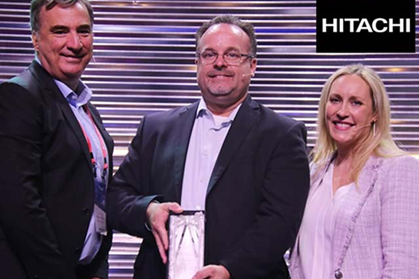 Three people in business attire smiling; middle person holding an award. Hitachi logo in the top right corner.