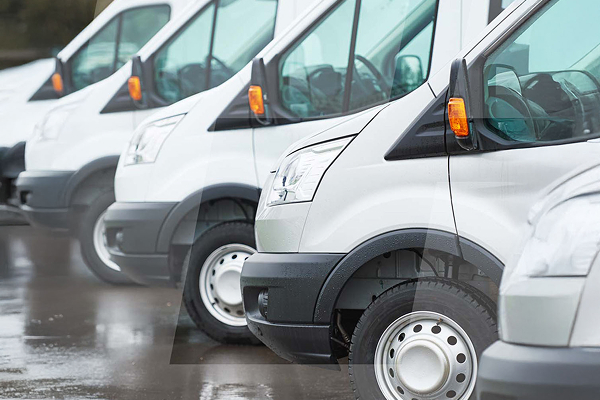 Row of white delivery vans parked in a lot, viewed from the front side, on a wet pavement.