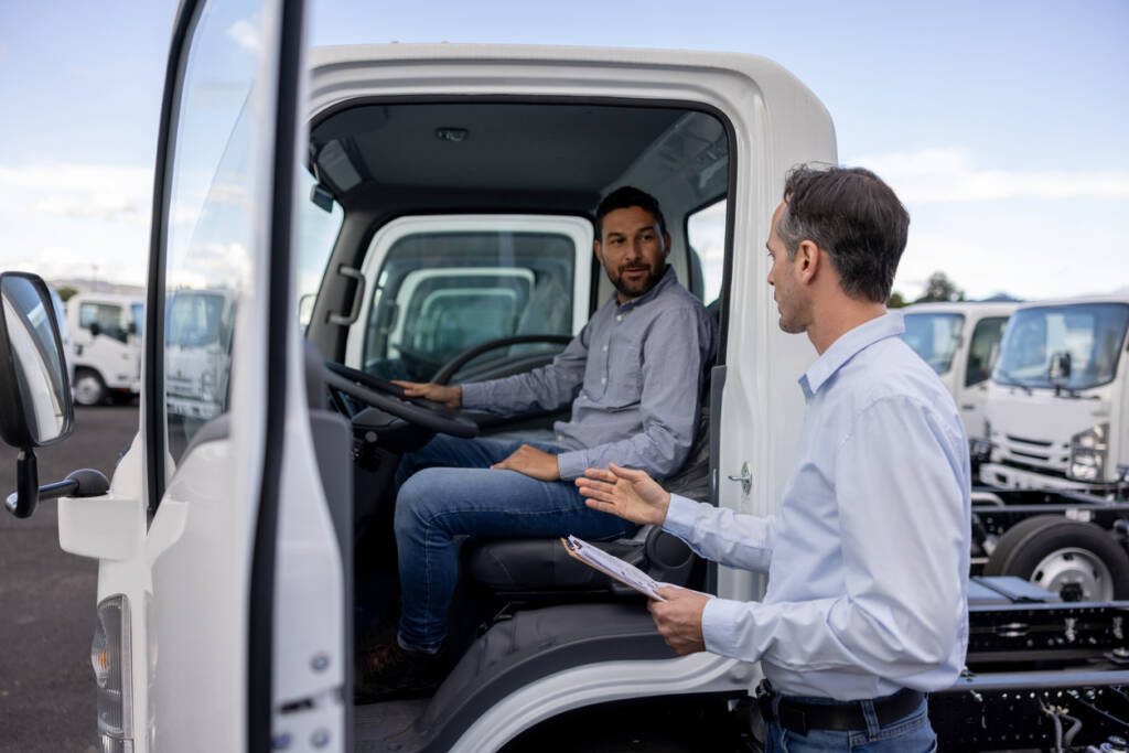 Person sitting in a truck cab talking to another person standing outside with a clipboard in a vehicle lot.