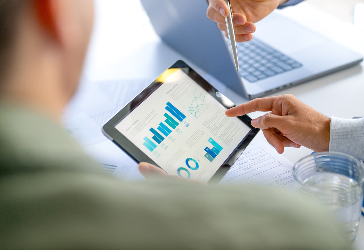 Two people discuss data on a tablet with charts and graphs, sitting at a desk with a laptop.