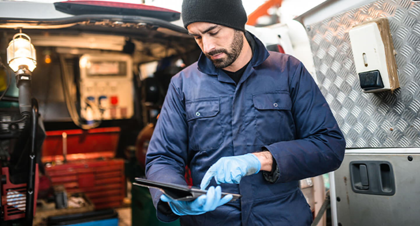 Technician in a blue jacket and beanie, using a tablet near a work van with tools and equipment inside.