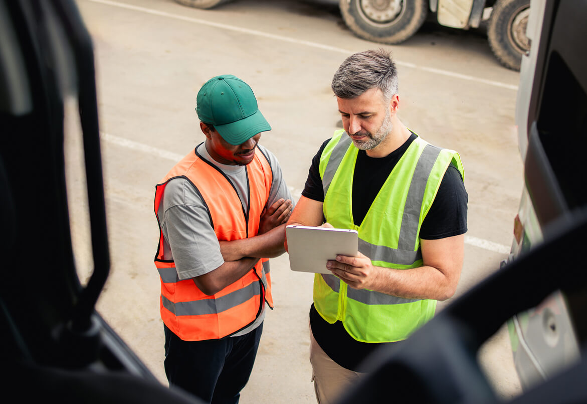 Two workers in reflective vests consult a clipboard near trucks on a paved surface.
