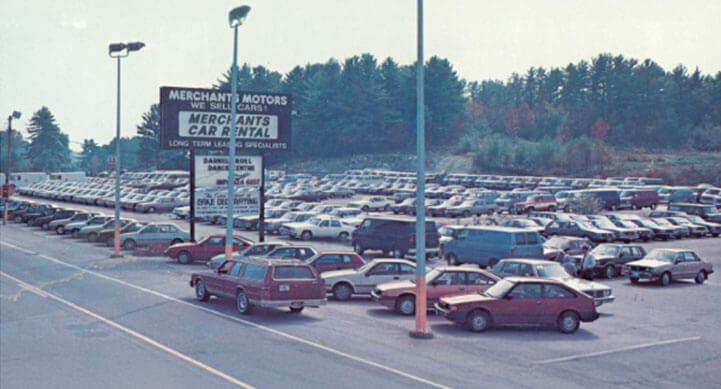 Large parking lot with rows of cars, a sign for Merchants Car Rental, and trees in the background.