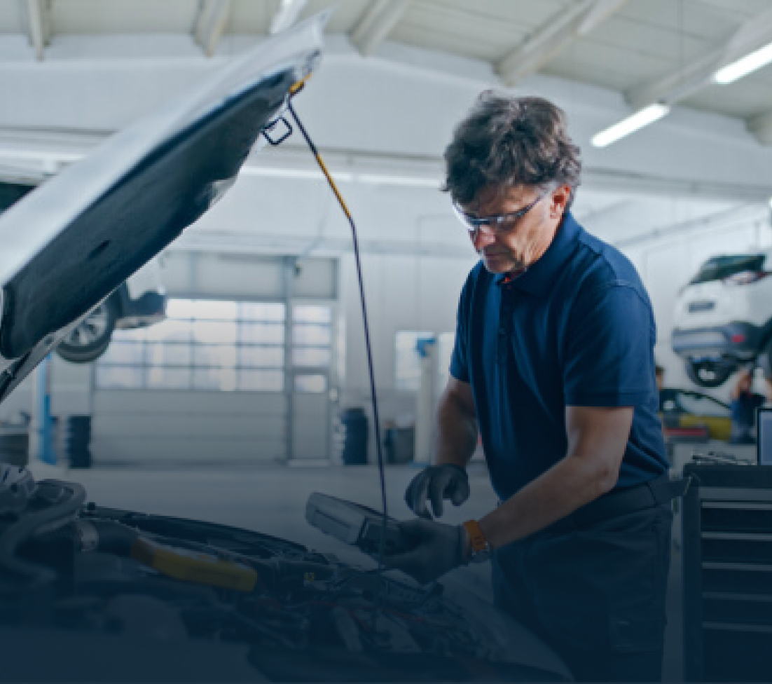 Mechanic wearing gloves and safety glasses, working under a car hood in a garage.