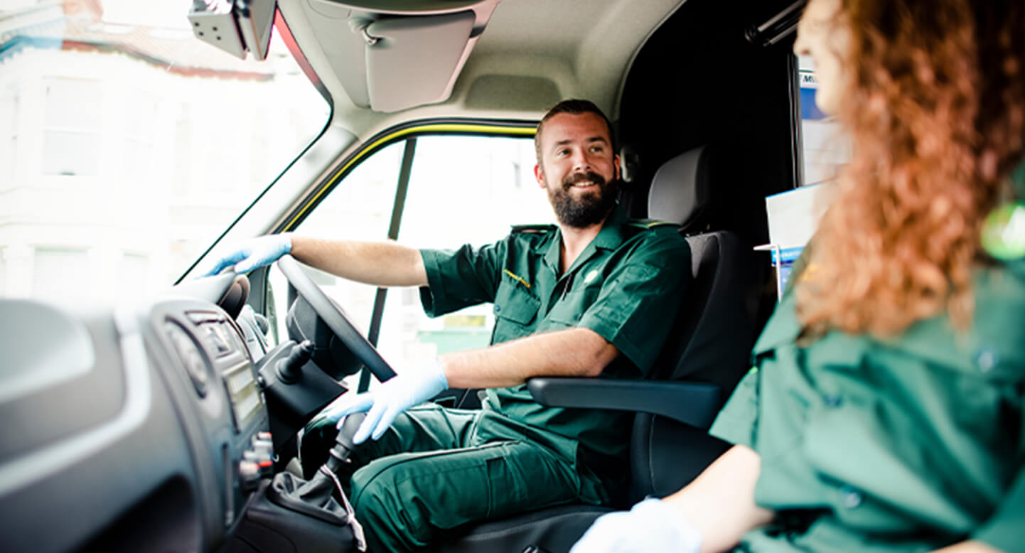 Two paramedics in green uniforms sit in an ambulance, smiling at each other.