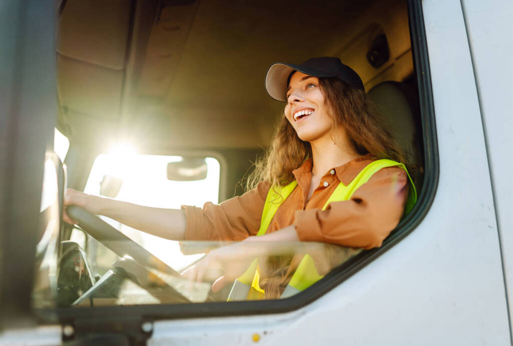 Smiling woman in a cap and safety vest driving a truck, with sunlight streaming in through the window.