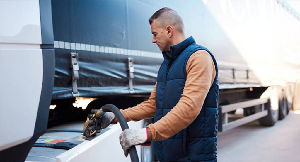 Man in a blue vest refueling a large truck with a nozzle, wearing gloves.
