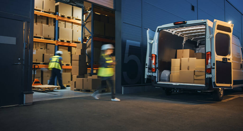 Workers load boxes into a white van at a warehouse loading dock at night.