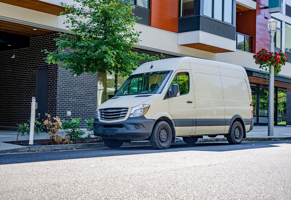 White delivery van parked on a street in front of modern buildings with trees and a hanging flower basket nearby.