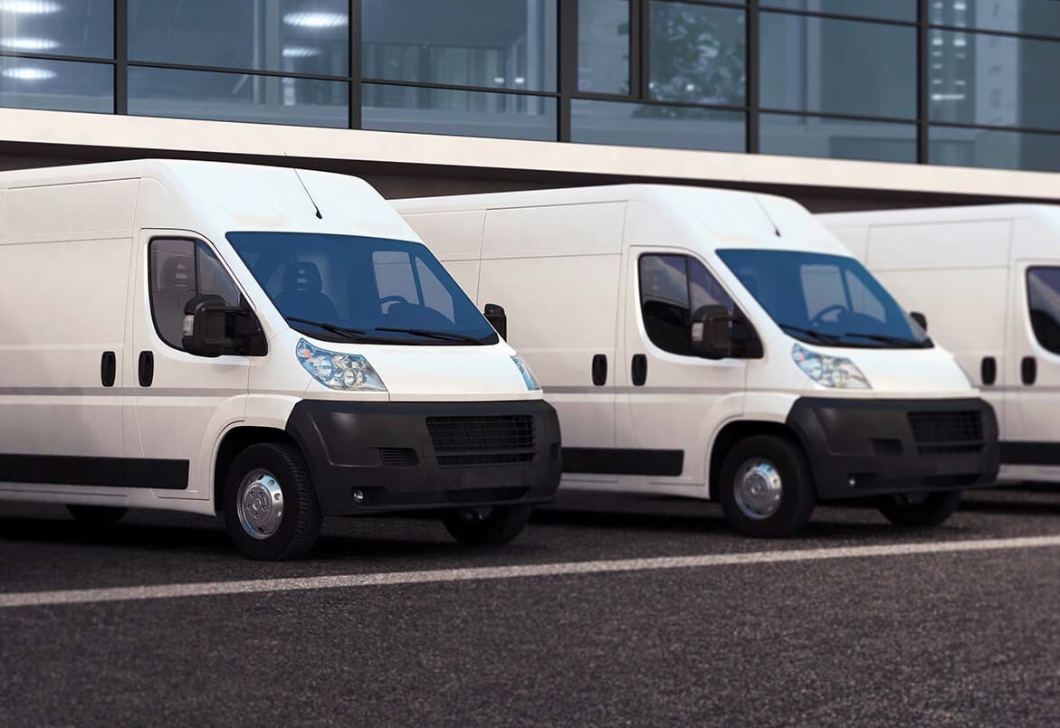 Three white delivery vans parked in a row outside a modern glass building on an asphalt surface.