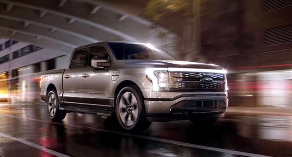 Silver pickup truck driving on a wet urban street at night with blurred city lights in the background.