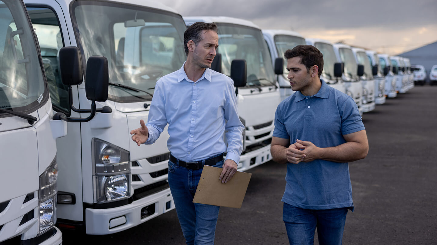 Two men talking and walking beside a row of white trucks in a parking lot. One holds a clipboard.