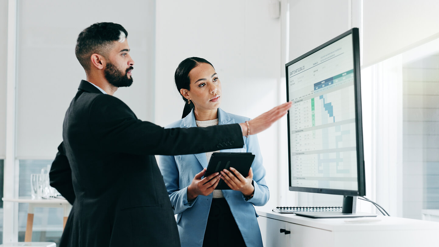 Two professionals analyzing data on a large monitor, with one holding a tablet and pointing at the screen.